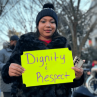 Black woman in black knit cap and coat standing outside in a protest holding a bright yellow handwritten sign that reads Dignity and Respect.