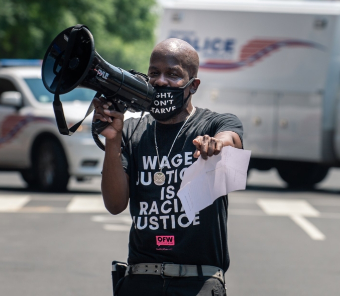 A Black man points directly at the photographer at an action. He is holding a bullhorn to the side and is wearing a mask that reads, "Fight, Don't Starve," and is wearing a shirt that reads "Wage Justice is Racial Justice."