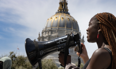 A black worker-organizer speaks into a black bullhorn at an action.