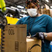 Young black woman with headwrap and facial mask packing a cardboard Amazon box inside a warehouse.