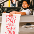 A man of color holding a sign that reads "FAIR PAY AND SAFE JOBS" while sitting in the driver's seat of a car, with a focused expression on their face.