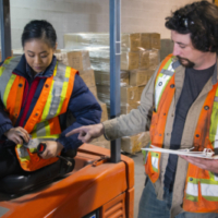 An industrial warehouse workplace safety topic. A safety supervisor or manager training a new employee on forklift safety. The trainer is holding a clipboard and explaining seat belt use to a younger female trainee.