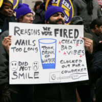 A worker at a City Hall action holds a sign that says "Reasons We Were Fired: Nails Too Long; Created Bad Vibes; Did Not Smile; Took Drink of Water; Clocked Out For A Coworker."