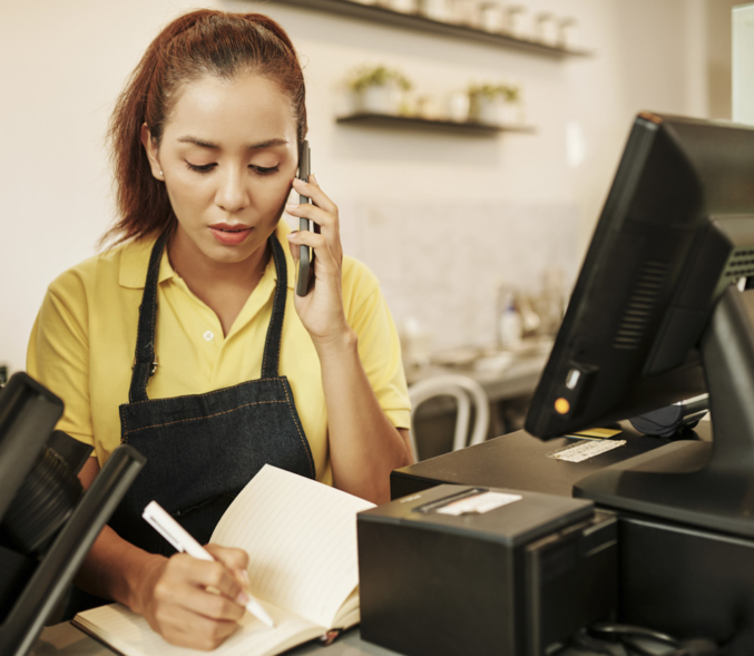 A woman at a workplace on the phone in front of a computer, taking down a message.