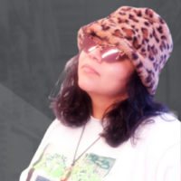 Image of Latinx woman with shoulder length brown hair wearing a bucket hat with animal print and sunglasses looking upwards.