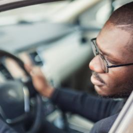 A Black man in a car looks out to his left-side mirror.