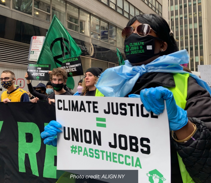 A Black woman wearing a mask and rubber gloves, leads a march holding a sign that reads: "Cimate Justice = Union Jobs, #PasstheCCIA" photo credit: Align NY