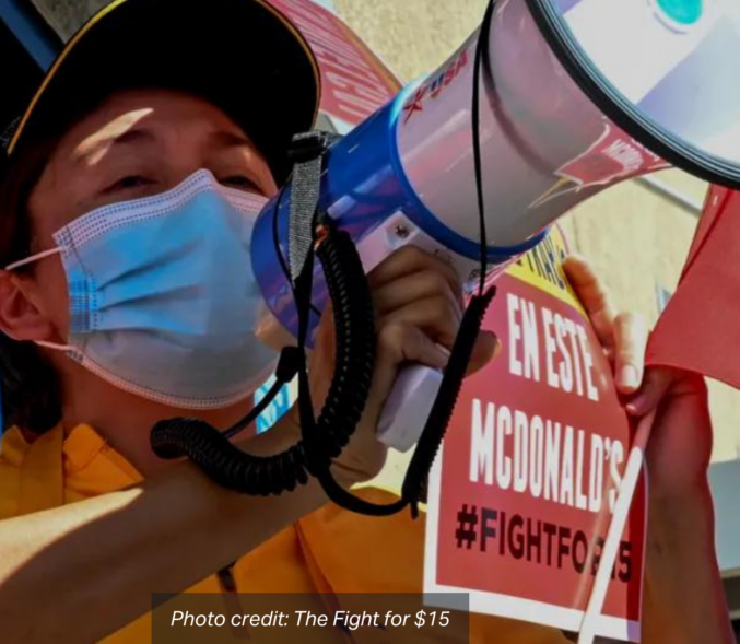 A worker wearing a McDonald's uniform and surgical mask speaks into a bullhorn in front of a poster that reads: "A History of Violence En Este McDonald's #Fightfor15". photo credit: The Fight for $15