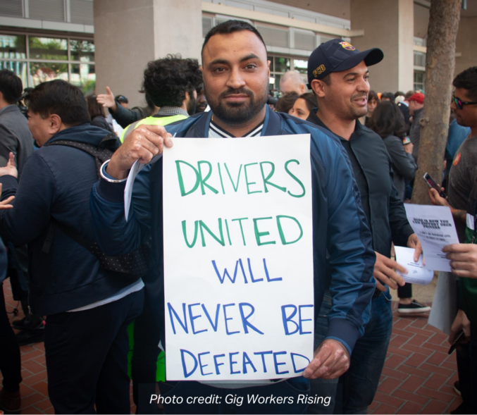 A man of Color holds a sign that reads: "Drivers United will Never be Defeated". photo credit: Gig Workers Rising