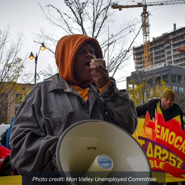 Frances Holmes, wearing an orange hooded sweatshirt and grey jacket, speaks into a bullhorn at a rally. photo credit: Mon Valley Unemployed Committee