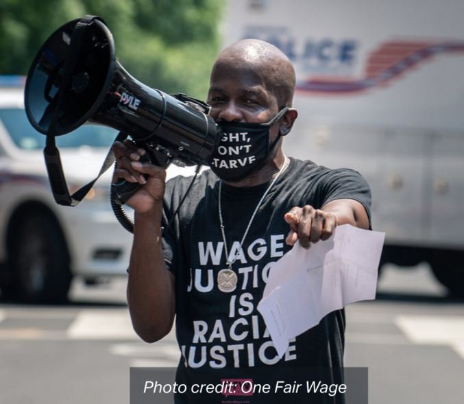 A Black man addresses a rally with a bullhorn, wearing a black mask that reads: "Fight, Don't Starve", and a shirt that reads: "Wage Justice is Racial Justice". photo credit: One Fair Wage