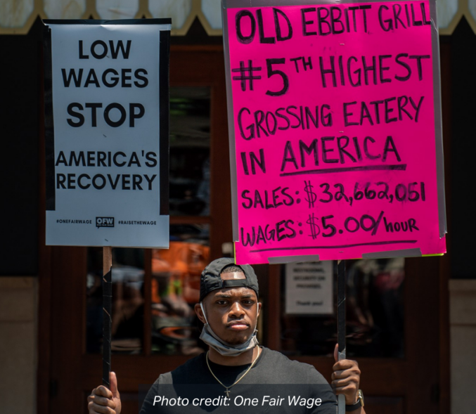 A Black man holds two signs that read: "Low Wages Stop America's Recovery", and "Old Ebbit Grill #5th highest grossing eatery in America. Sales: $32,662,051 Wages: $5.00/hour". photo credit: One Fair Wage