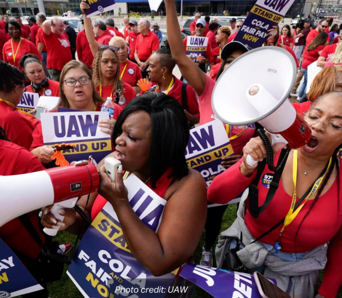 Two Black woman speak into bullhorns at a United Auto Workers Rally. The crowd wears red shirts and hold signs that read: "Stand Up COLA and Fair Pay Now". photo credit: UAW