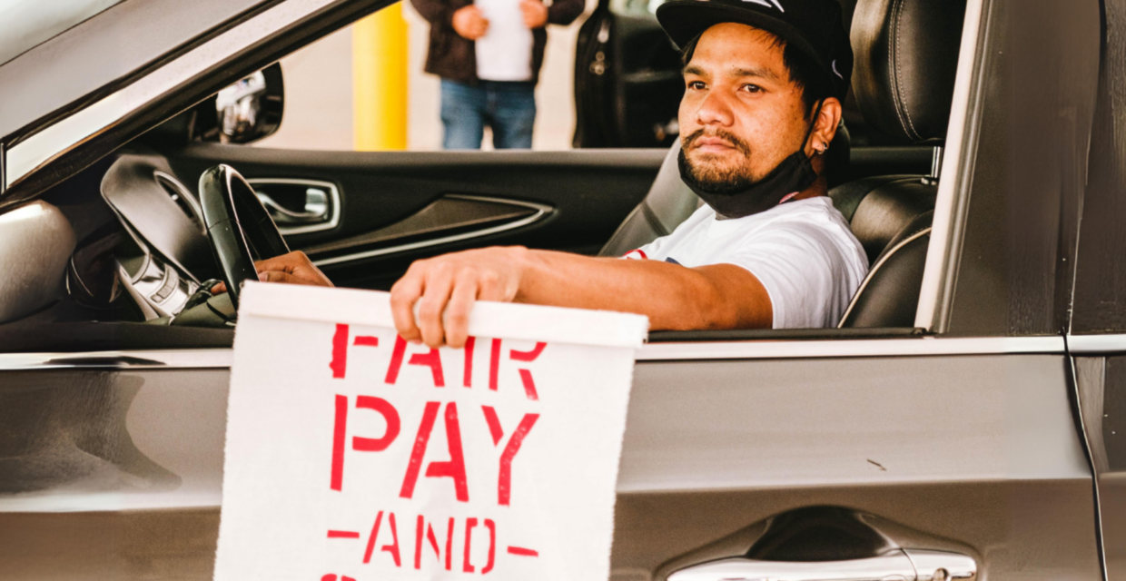 Ridehail driver sits in his car holding a sign that says "Fair Pay and Safe Jobs".