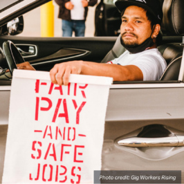 Ridehail driver sits in his car holding a sign that says "Fair Pay and Safe Jobs".