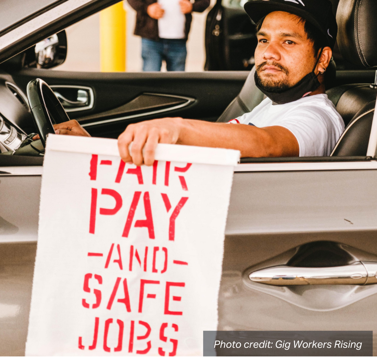 Ridehail driver sits in his car holding a sign that says "Fair Pay and Safe Jobs".