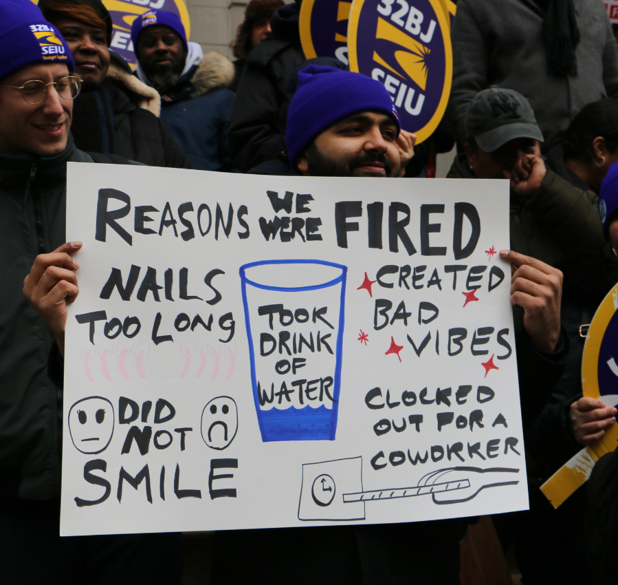 SEIU members attend a rally holding a sign that reads "Reasons we were fired - Nails too long, Created bad vibes, did not smile, clocked out for a coworker, took drink of water"