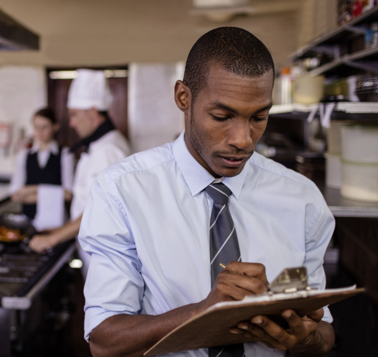 A young Black man writes on a clipboard with coworkers in a kitchen setting